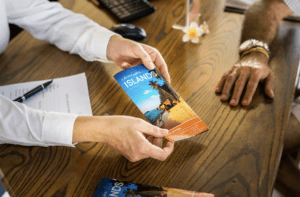 Person handing a colorful brochure titled "ISLANDS" to another person at a wooden table, highlighting brochure design and marketing in a business setting.