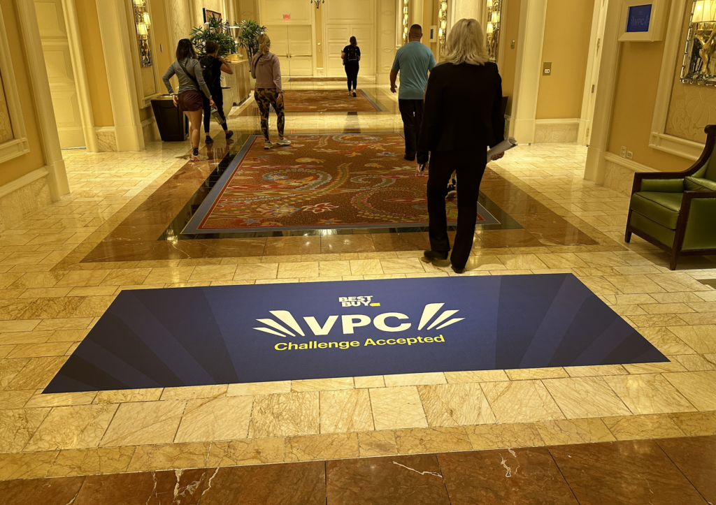 Floor graphic displaying "VPC Challenge Accepted" in a busy hallway, with people walking by and decorative elements in the background, emphasizing creative marketing solutions.