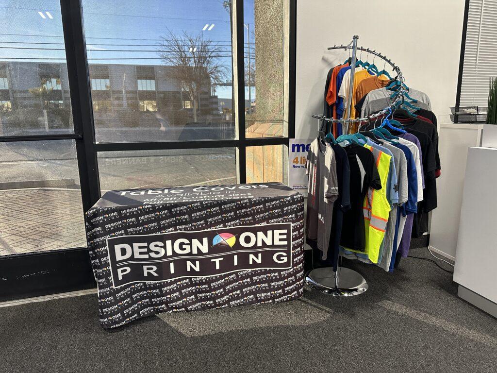 Table cover featuring "Design One Printing" branding, displayed in a printing service environment alongside a rack of colorful shirts, emphasizing promotional materials for events and marketing.