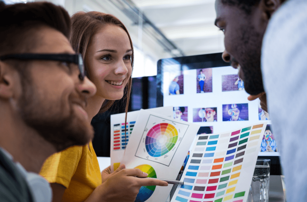 Three individuals discussing color palettes and design options, with color swatches and a computer displaying images in a creative workspace, reflecting collaboration in graphic design for convention printing.