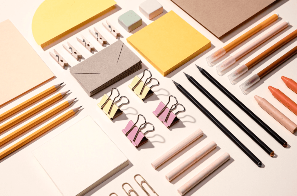 Assorted stationery items including pencils, markers, sticky notes, paper clips, binder clips, and envelopes, arranged on a flat surface, emphasizing office supplies and promotional products for printing services.