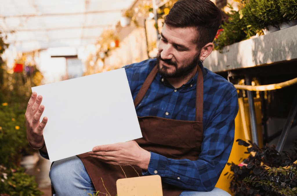 Man in plaid shirt and apron holding blank white sign in a garden setting, illustrating the concept of customizable printing services for promotional materials.