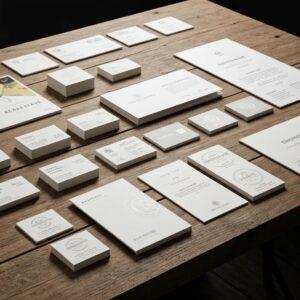 Collection of various printed stationery items including business cards, letterheads, and envelopes displayed on a wooden surface, showcasing design elements relevant to branding and marketing materials.